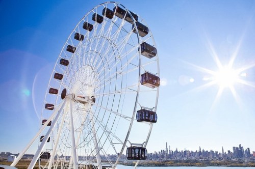 A large white Ferris wheel against a clear blue sky, with a city skyline in the background and the sun shining brightly.