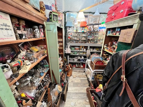 A narrow aisle filled with various vintage items and collectibles on shelves and tables in a cluttered shop.