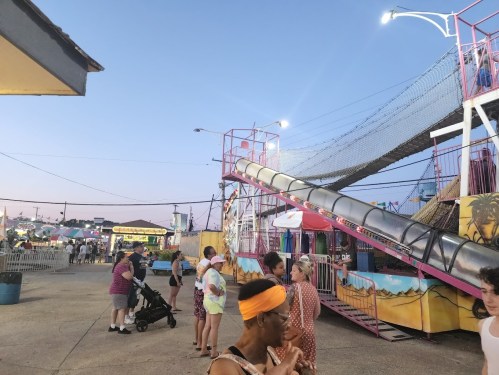 A lively carnival scene at dusk, featuring a slide, colorful stalls, and people enjoying the festivities.