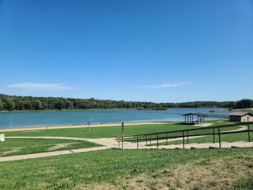 A serene lake view with a sandy beach, green grass, and a clear blue sky in the background.