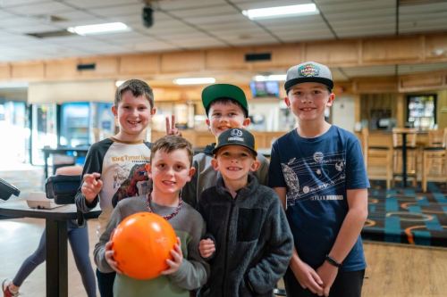 Five smiling children pose together at a bowling alley, holding an orange bowling ball.