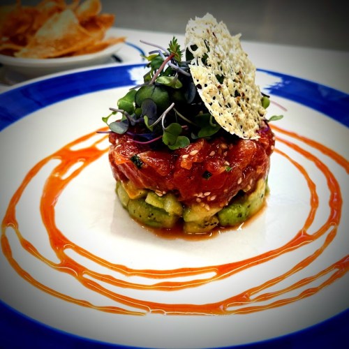 A colorful dish featuring layered tuna tartare, avocado, microgreens, and a decorative sesame crisp on a blue plate.