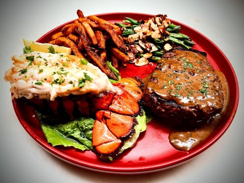 A vibrant plate featuring a lobster tail, steak with gravy, sweet potato fries, and green beans with almonds.