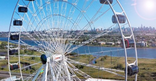 Aerial view of a large Ferris wheel with city skyline and river in the background on a clear day.