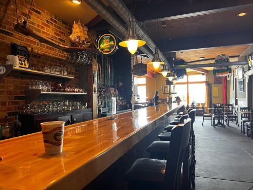A spacious bar with a wooden counter, stools, and a brick wall, featuring drinks and a few patrons in a cozy setting.