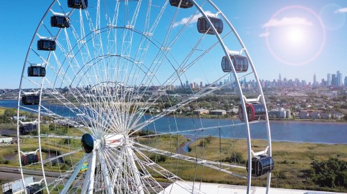 Aerial view of a large Ferris wheel by a river, with a city skyline in the background under a bright sun.