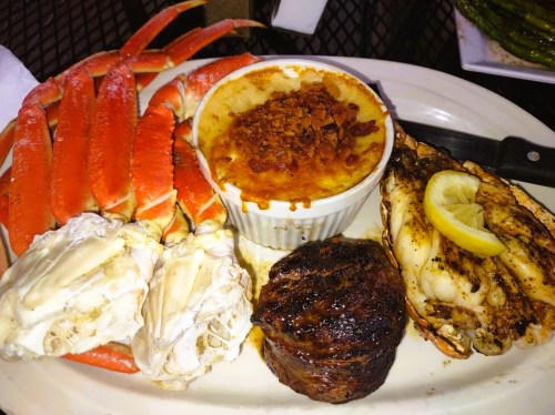 A plate featuring crab legs, a baked dish with bacon, and grilled seafood alongside a steak, garnished with lemon.