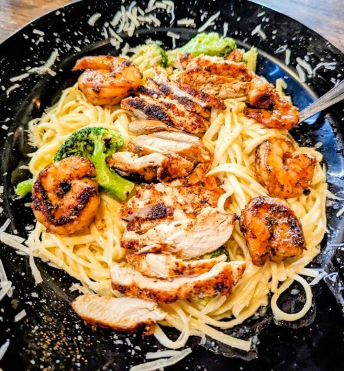 A plate of pasta topped with grilled chicken, shrimp, and broccoli, garnished with grated cheese.