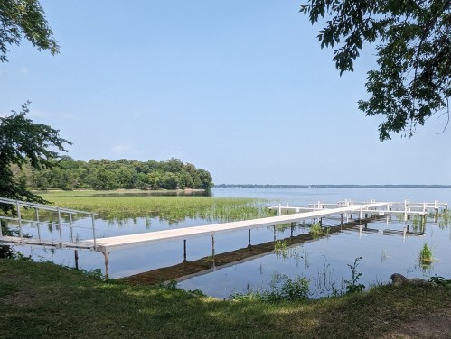A serene lake view featuring a wooden dock extending into calm waters, surrounded by lush greenery and blue skies.