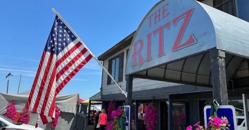A building with a sign reading "The Ritz," featuring a large American flag and colorful flowers in front.