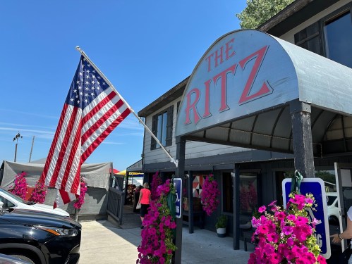 Exterior of The Ritz with a large American flag and vibrant pink flowers adorning the entrance on a sunny day.