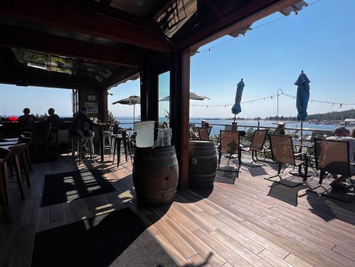 A cozy waterfront restaurant interior with wooden floors, barrels, and patrons enjoying the view of the water.