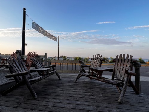 Adirondack chairs on a wooden deck overlooking a calm waterfront at sunset, with a banner in the background.