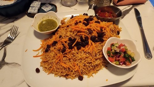 A large plate of rice topped with carrots and raisins, accompanied by green sauce, chutney, and a fresh salad.