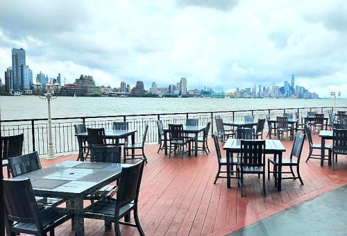 Outdoor dining area with wooden tables overlooking a river and city skyline under a cloudy sky.