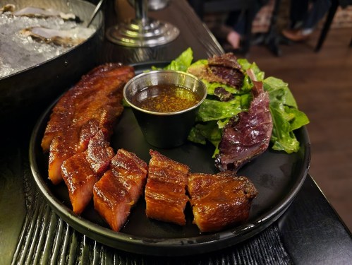 A black plate with sliced grilled meat, a small bowl of sauce, and a side of mixed greens.