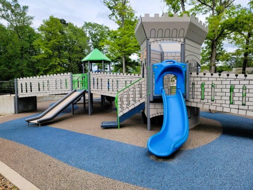 Colorful playground with a castle theme, featuring slides and climbing structures, surrounded by green trees.