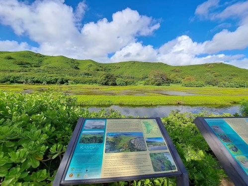Lush green hills under a blue sky, with informational signs in the foreground near a tranquil wetland area.