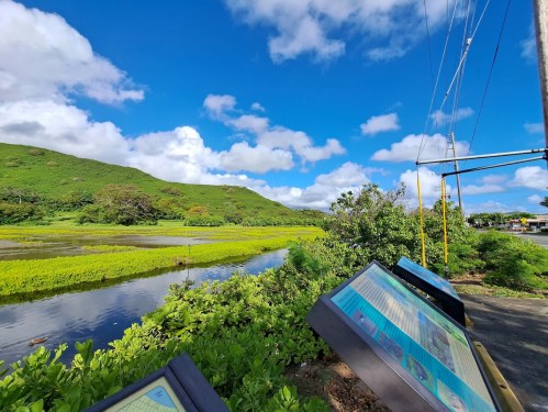 A scenic view of lush green hills and a calm waterway, with informational signs in the foreground under a blue sky.