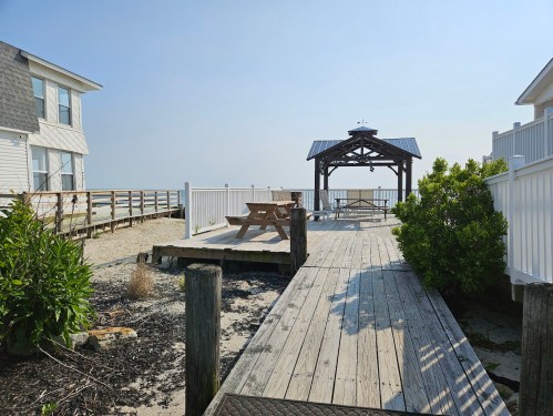A wooden walkway leads to a gazebo by the beach, surrounded by white fences and a picnic table nearby.