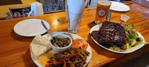 A wooden table with two plates of food: one with grilled meat, vegetables, and a tortilla; the other with ribs and salad.