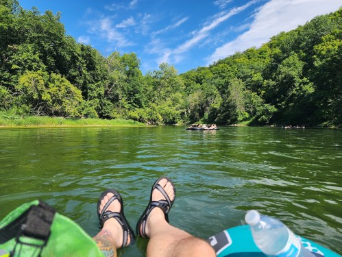 Person's legs in sandals resting on a float in a calm river, surrounded by lush green trees and a clear blue sky.