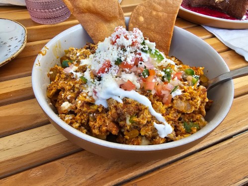A bowl of chilaquiles topped with cheese, sour cream, tomatoes, and tortilla chips, served on a wooden table.