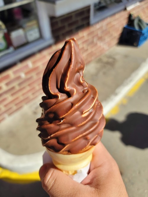 A hand holding a chocolate soft serve ice cream cone with a swirled top against a brick background.