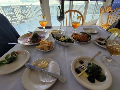 A dining table with various dishes, drinks, and a view of the water in the background.