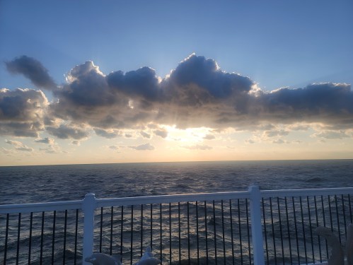 Sunset over the ocean, with clouds illuminated by golden light, viewed from a pier with a white railing.