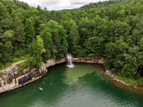 Aerial view of a lush green forest surrounding a serene waterfall and calm water, with kayakers enjoying the scene.