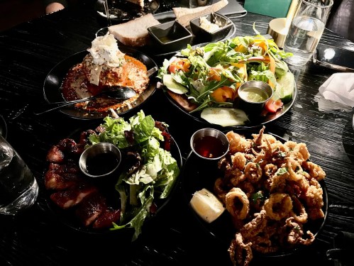 A table with four plates of food: a salad, fried calamari, grilled meat, and a dessert topped with whipped cream.