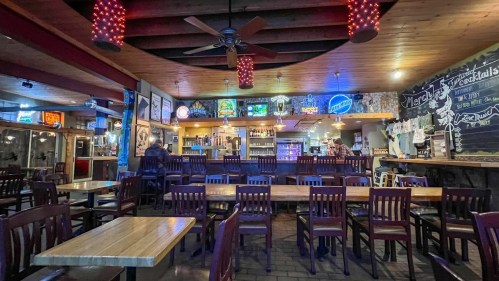 Interior of a cozy bar with wooden tables, chairs, a long bar, and colorful neon signs on the walls.