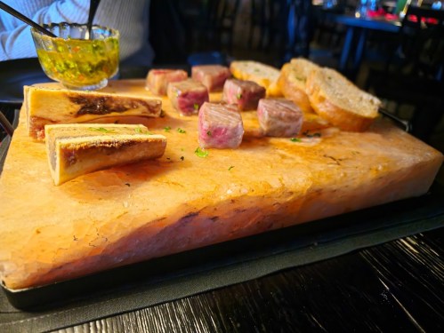 A wooden platter with grilled meats, bone marrow, and slices of bread, accompanied by a small bowl of sauce.