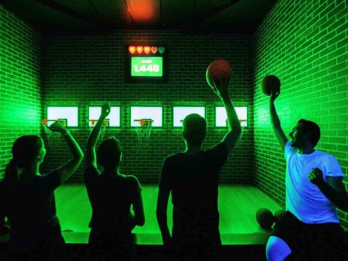 Four people play basketball in a neon-lit arcade, raising their balls in excitement with a scoreboard in the background.