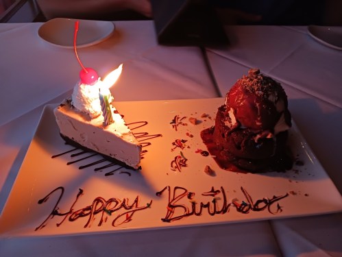A birthday dessert plate featuring cheesecake and a chocolate cake, topped with whipped cream, a cherry, and a candle.