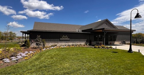 A modern building with a dark exterior, surrounded by green grass and colorful flowers under a blue sky with clouds.