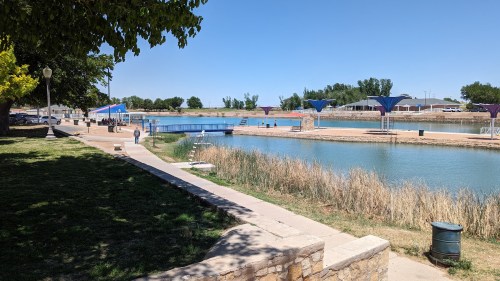 A serene park with a pathway alongside a calm waterway, featuring docks and greenery under a clear blue sky.