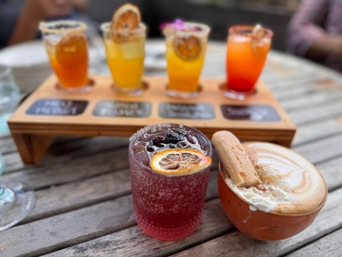 A wooden tray holds five colorful cocktails, with a red drink and a dessert on the table, all set outdoors.
