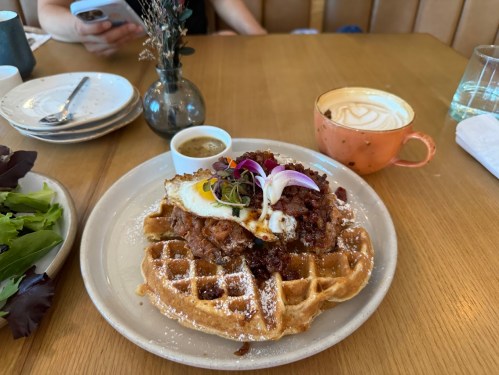 A plate of waffles topped with fried chicken, an egg, and garnished with herbs, alongside a small sauce and a coffee.