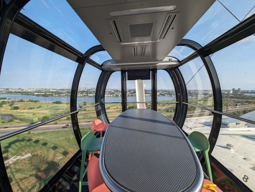 A modern glass dining pod with a panoramic view of a landscape and water, featuring a sleek table and colorful chairs.