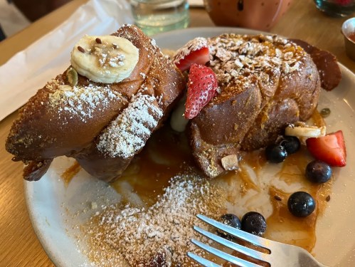 A plate of French toast topped with banana, strawberries, blueberries, and powdered sugar, drizzled with syrup.
