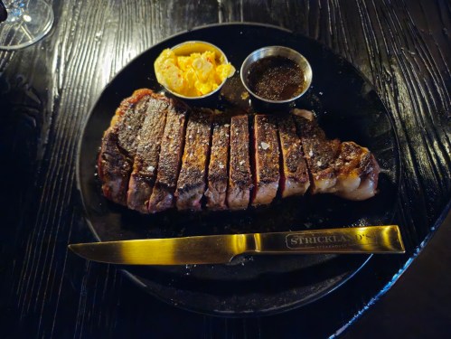 A perfectly grilled steak, sliced and served on a black plate with sides of macaroni and dipping sauce.