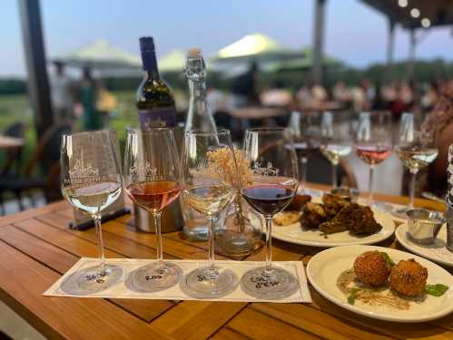 A wooden table with wine glasses, a bottle, and plates of food, set outdoors with a scenic background.