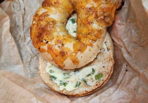 A toasted bagel topped with melted cheese, resting on a slice spread with cream cheese and herbs.