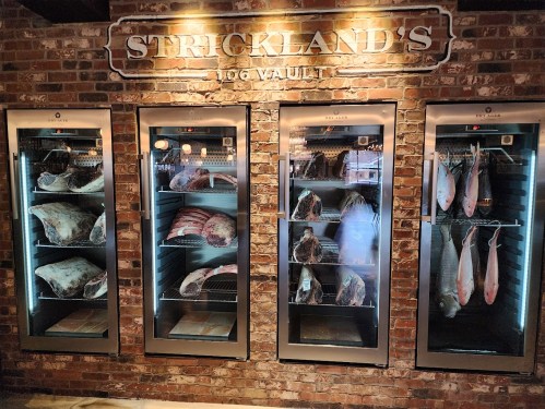 A display of meat and fish in glass refrigerators against a brick wall, labeled "Strickland's Meat Vault."