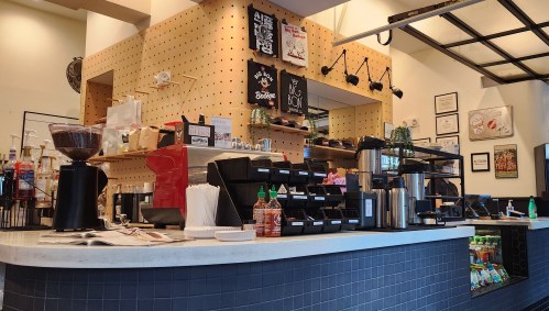A modern café counter with coffee machines, condiments, and a menu board, featuring a warm, inviting design.