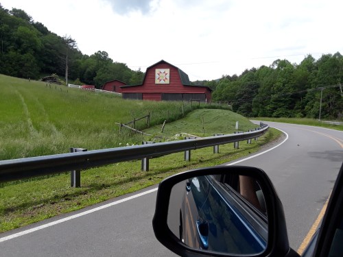 A red barn with a sun emblem sits beside a winding road, surrounded by green fields and trees.