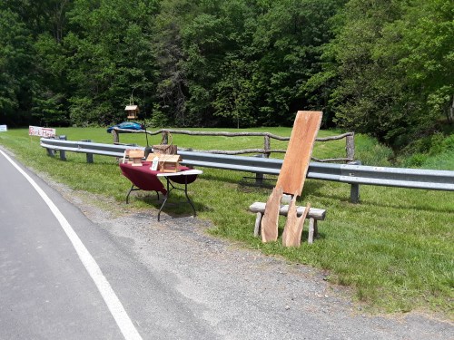A roadside stand with wooden items for sale, set against a green landscape and guardrail.