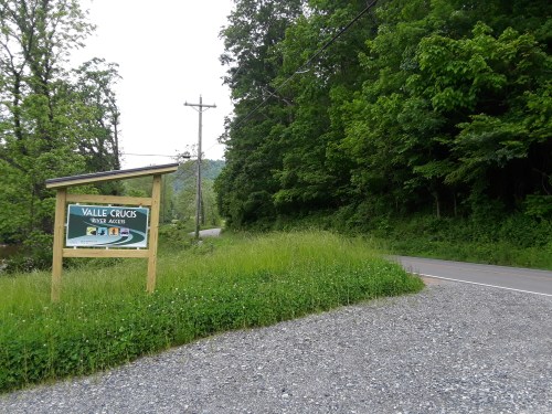 A sign for Valle Crucis, surrounded by greenery, with a gravel path leading to a road in the background.